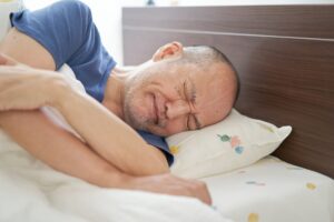 Man with gray beard in bed clenching eyes and teeth